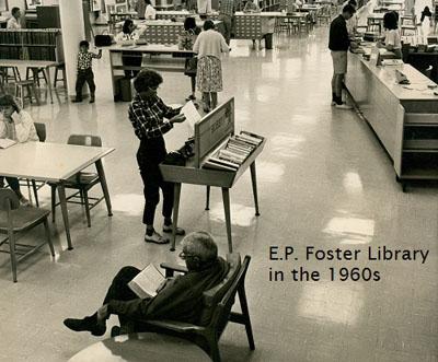 Black and white photo of the E.P. Foster Library in the 1960s. Shows card catalogs, a long reference desk, book stacks, and tables. About 14 people are shown in the photo. 
