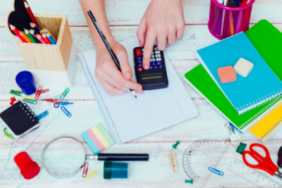 Photo of hands using a pencil and calculator on a white desk surrounded by colorful study supplies.