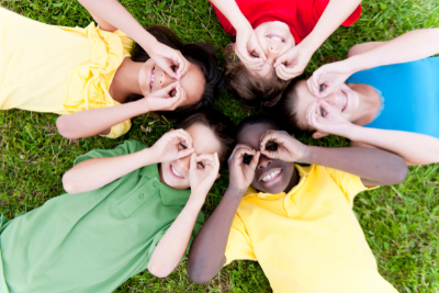 Smiling children laying on grass, heads toward center, holding hands over their eyes like binoculars.