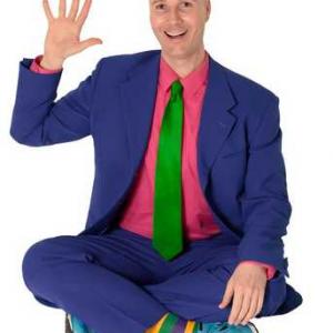 Christopher in a purple suit, hot pink shirt, green tie, yellow/purple/green striped socks, and blue sneakers. He is waving while sitting cross-legged and smiling.
